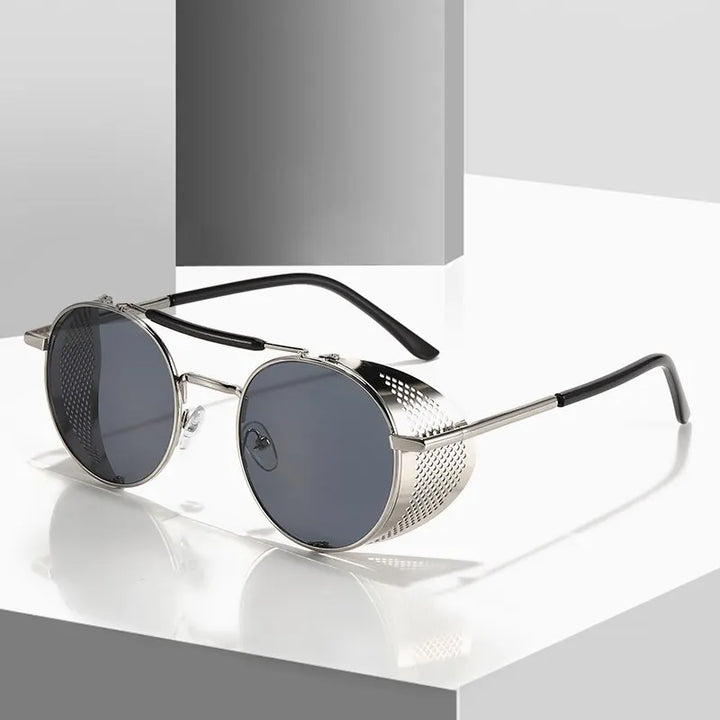 Round Steampunk sunglasses with dark tinted lenses and silver mesh side shields rest on a glossy, white geometric surface. These windproof mesh sunglasses feature a modern design with black ear tips and a striking metallic finish.