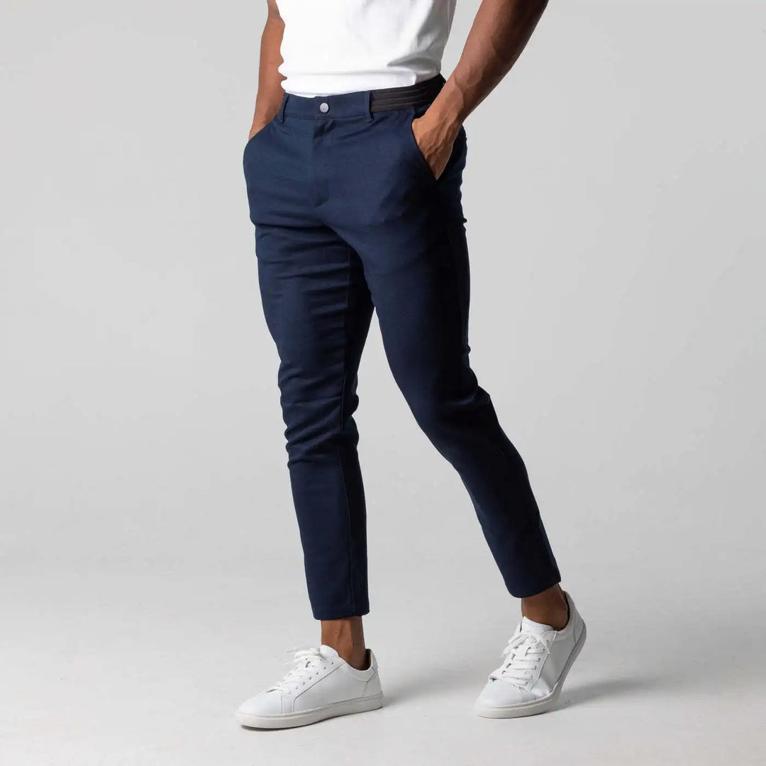 A person wearing a white t-shirt, navy blue slim fit ankle length cropped pants with white sneakers stands with hands in pockets against a plain gray background.