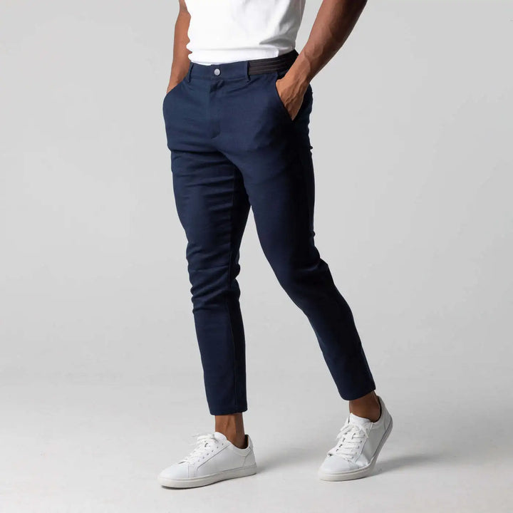 A person wearing a white t-shirt, navy blue slim fit ankle length cropped pants with white sneakers stands with hands in pockets against a plain gray background.
