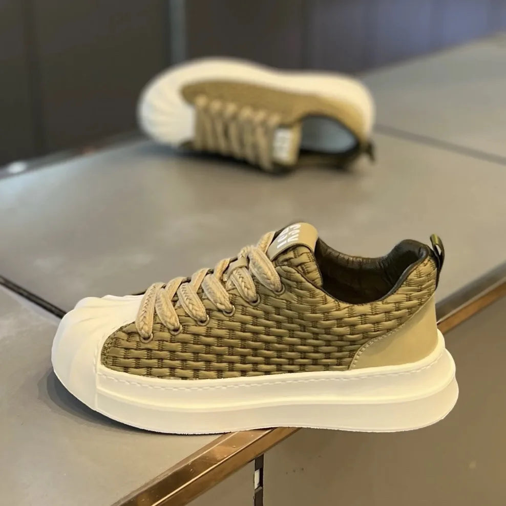 A stylish lightweight shoe with a woven olive-green upper, cushioned white chunky sole, and beige toe cap is displayed on a counter. Another matching sneaker is blurred in the background.