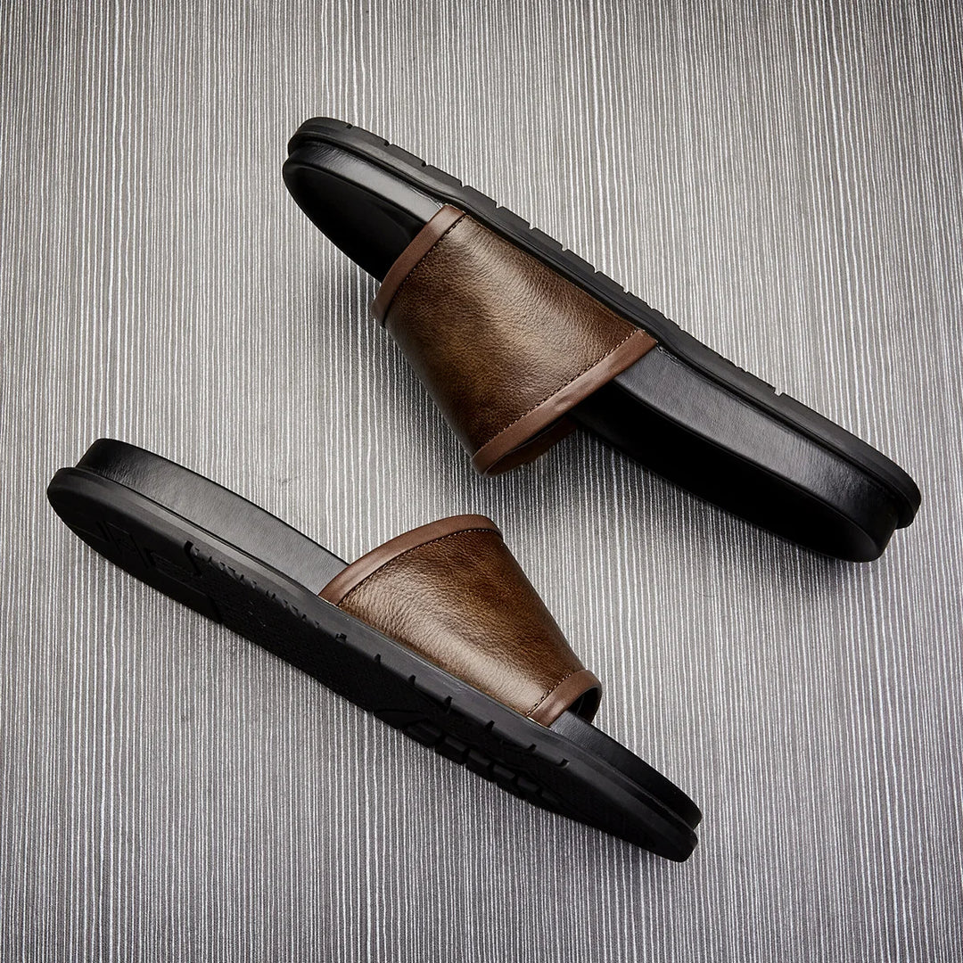 A pair of men's luxury sandals in brown and black with wide straps, positioned diagonally on a textured, light gray background.