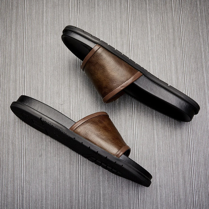 A pair of men's luxury sandals in brown and black with wide straps, positioned diagonally on a textured, light gray background.