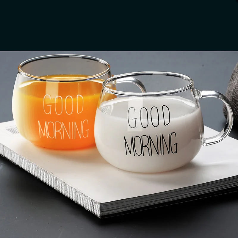 Two clear glass mugs featuring a clear design and “GOOD MORNING” print, one with orange juice and the other with milk, rest side by side on a closed white book against a dark background.