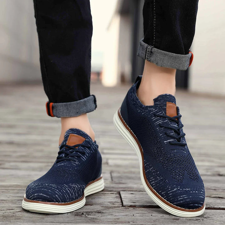A person wearing dark blue, lightweight sneakers with white soles and brown accents stands on a wooden surface, jeans rolled up at the ankles for a relaxed, casual style.