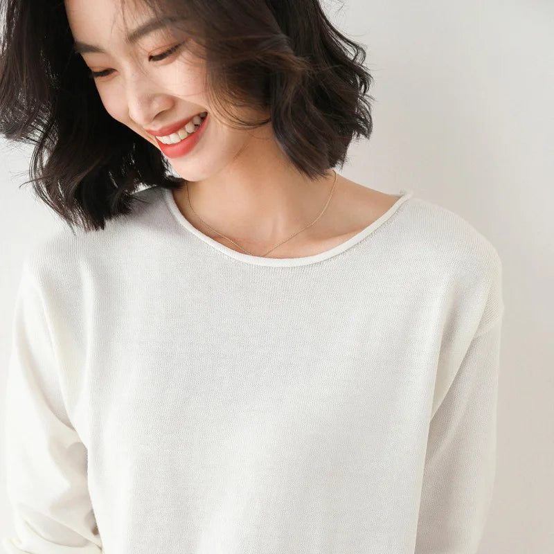 A woman with shoulder-length dark hair smiles while looking down. She is wearing a relaxed fit, light-colored sweater and a delicate necklace, standing against a neutral background.