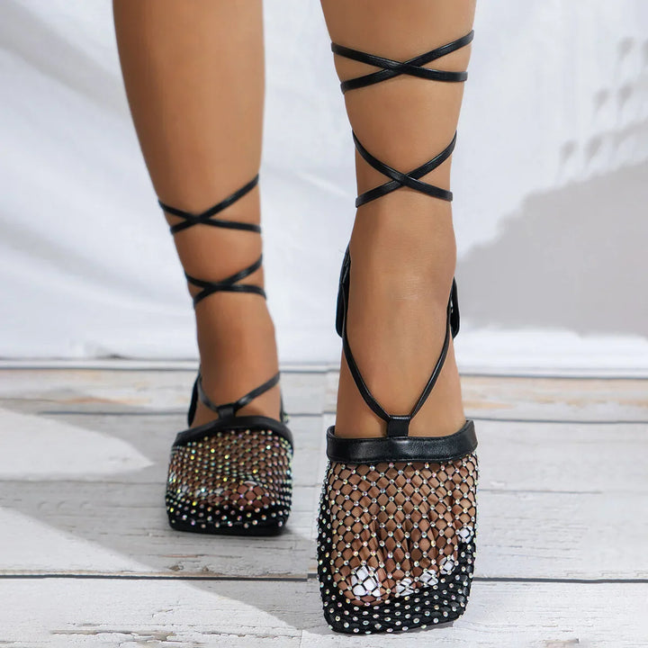 A person wearing black strappy sandals with a slip-resistant sole and glittery rhinestone mesh upper over the front, standing on white wooden flooring.