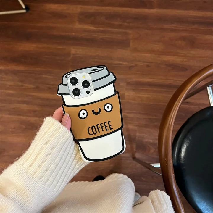 A hand in a cream-colored sweater holds a phone with a cute 3D coffee cup-shaped case, featuring a smiling face and the word COFFEE. Made from soft silicone for shockproof protection. The background shows a wooden floor and part of a chair.