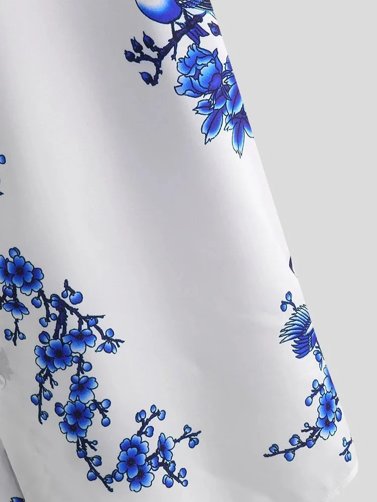 A close-up of a cotton-silk blend white fabric featuring a blue floral and bird print with intricate branches and blossoms, set against a plain gray background—a summer-ready choice.