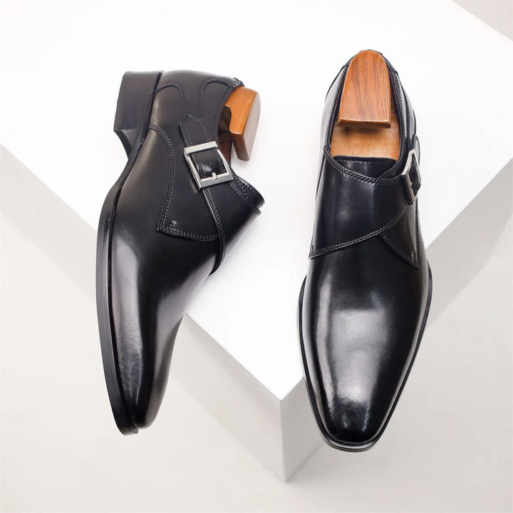 A pair of polished black Italian leather monk strap shoes with silver buckles, displayed on a white geometric surface. One handcrafted shoe is lying flat, while the other is propped up, both with wooden shoe trees inside.