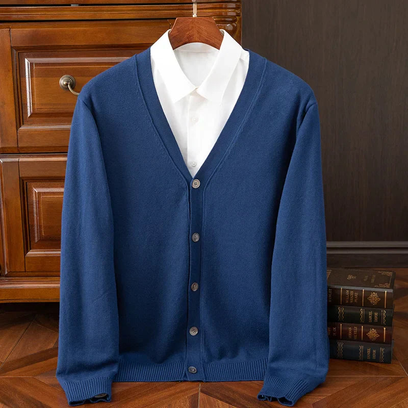 A blue button-up cardigan with a V-neckline and relaxed fit, layered over a white collared shirt, is displayed on a hanger in front of a wooden dresser and some stacked books.