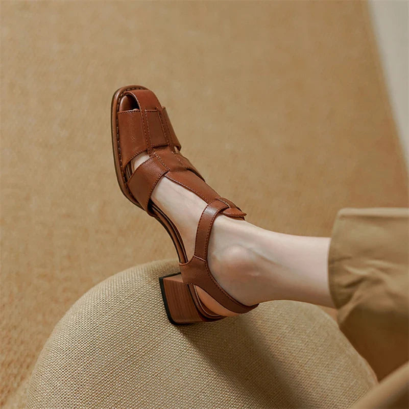 A close-up of a person’s foot wearing a brown, open-toe sandal with ergonomic arch support and a block heel. Crafted from Genuine Italian Leather, the sandal rests on a textured beige surface, paired with light brown pants.