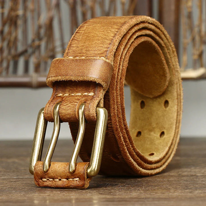 A rolled-up genuine leather belt with a double brass buckle rests on a wooden surface, showcasing heritage craftsmanship against a blurred rustic background.