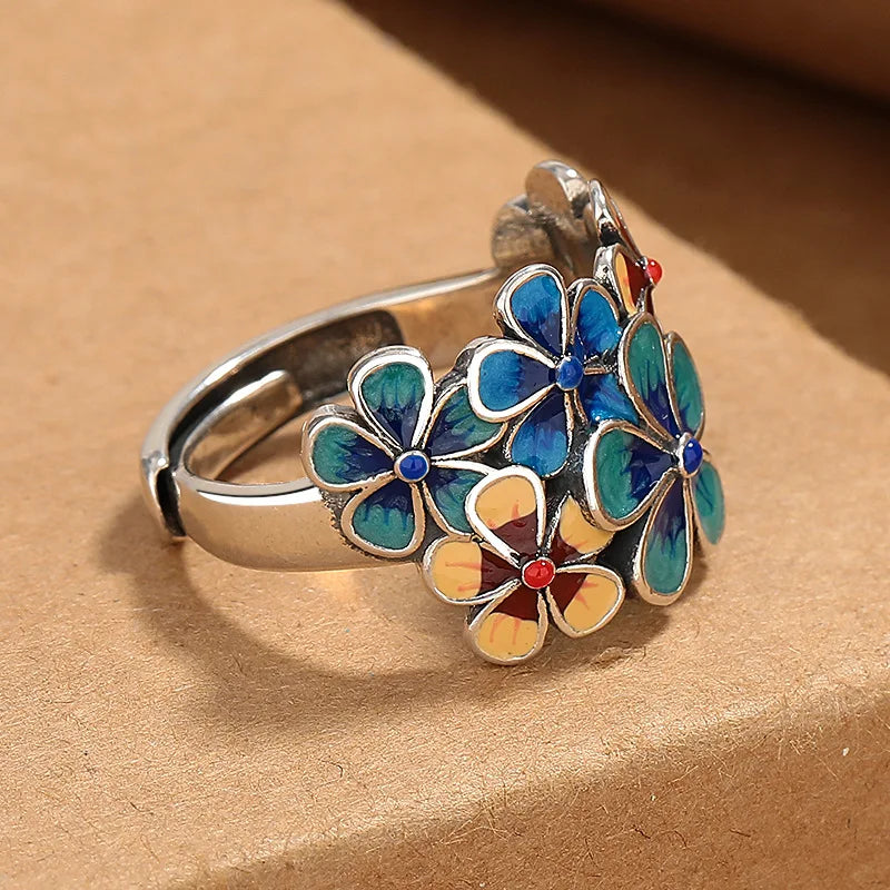 A silver ring with a cluster of colorful enamel flowers in blue, teal, yellow, and red, featuring vibrant colors reminiscent of stained glass, displayed on a brown textured surface.