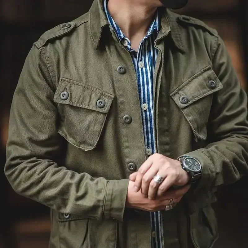 A person wearing a vintage military jacket in olive green over a blue-and-white striped shirt adjusts a black watch on their wrist; their head is out of frame.