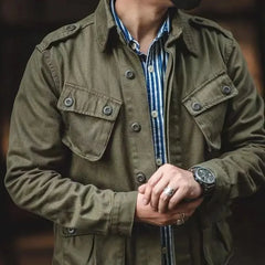 A person wearing a vintage military jacket in olive green over a blue-and-white striped shirt adjusts a black watch on their wrist; their head is out of frame.