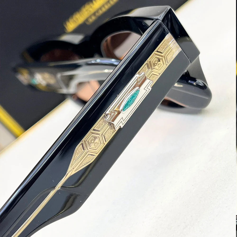 Close-up of stylish black vintage sunglasses with intricate gold and silver geometric detailing on the temple, featuring a small green accent in the center. The blurred background highlights the glasses design.