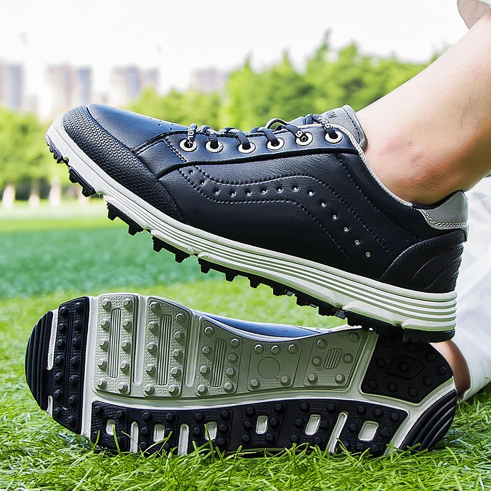 A close-up of a person wearing black waterproof golf shoes with white soles, standing on green grass. The advanced cleat technology and tread pattern are clearly visible. Trees and blurry buildings appear in the background.
