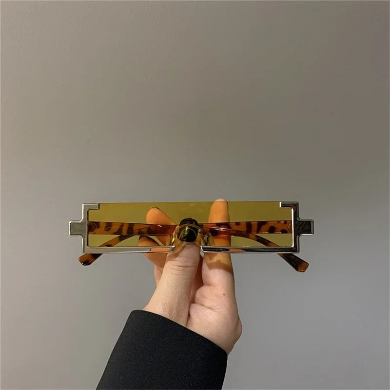 A hand holds a pair of rectangular, yellow-tinted sunglasses with unique geometric silver frames and tortoiseshell-patterned arms featuring scratch-resistant lenses, all set against a plain gray background.