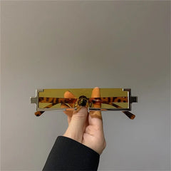 A hand holds a pair of rectangular, yellow-tinted sunglasses with unique geometric silver frames and tortoiseshell-patterned arms featuring scratch-resistant lenses, all set against a plain gray background.