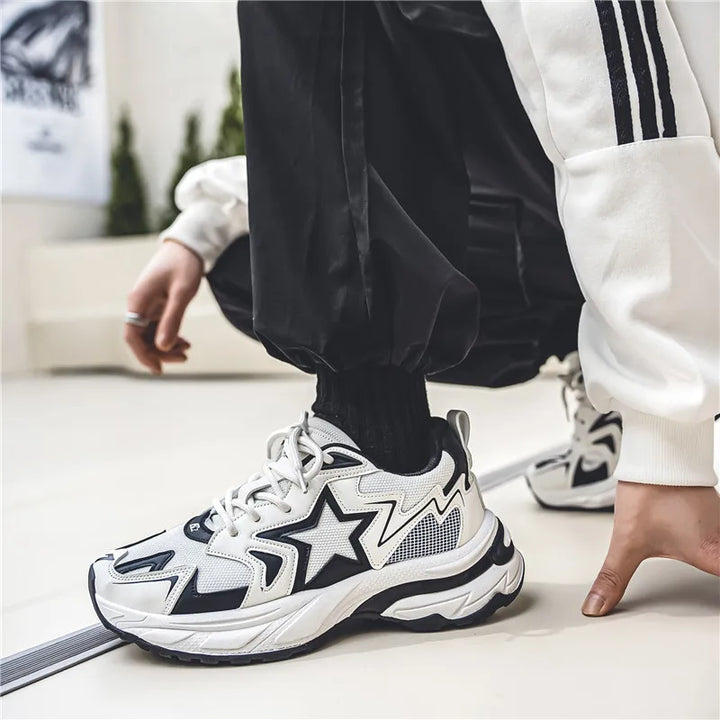 A person in black joggers and a white jacket crouches, wearing white sneakers with bold black star designs, a durable rubber outsole, and wavy patterns on the soles. The setting appears to be indoors.