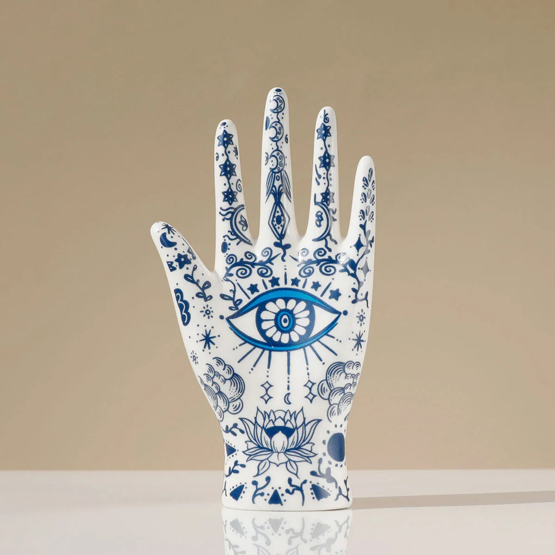 A white ceramic hand sculpture decorated with intricate blue designs, including an eye on the palm and a lotus flower near the base—perfect as a boho-chic home décor accent or unique ceramic jewelry stand with celestial and evil eye decor elements.