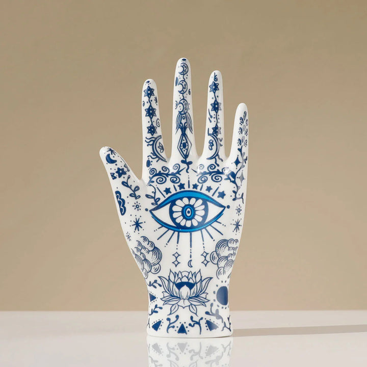 A white ceramic hand sculpture decorated with intricate blue designs, including an eye on the palm and a lotus flower near the base—perfect as a boho-chic home décor accent or unique ceramic jewelry stand with celestial and evil eye decor elements.