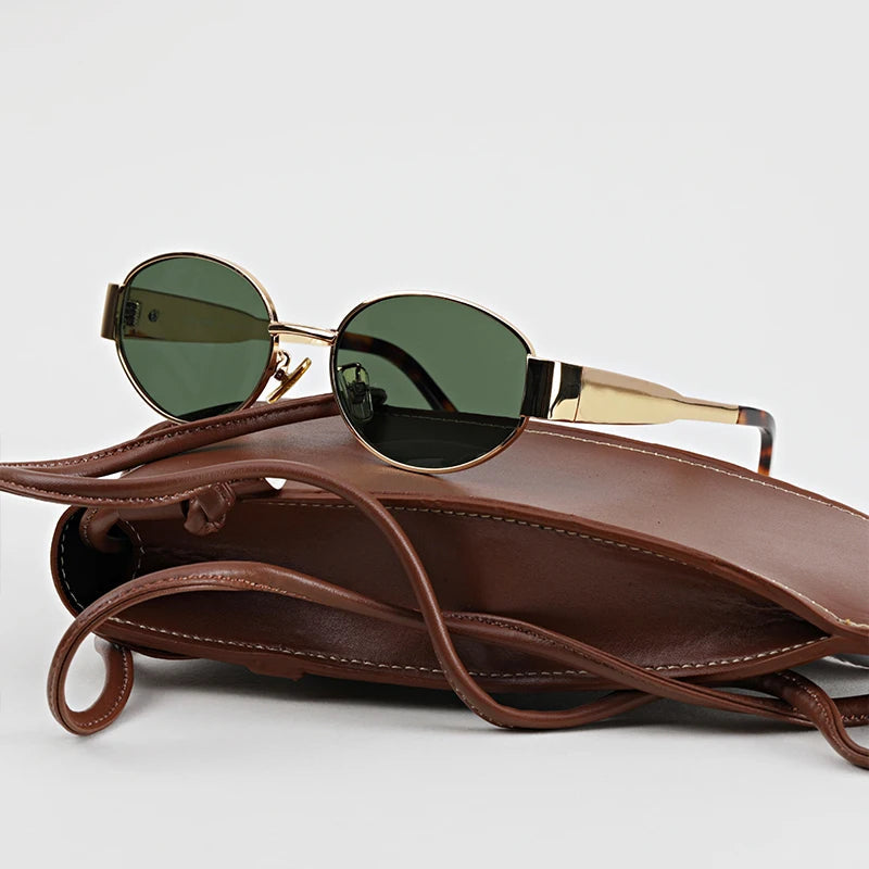 A pair of vintage-inspired, gold-framed oval green sunglasses with UV400 protection rests on a brown leather handbag with visible stitching, set against a plain light background.