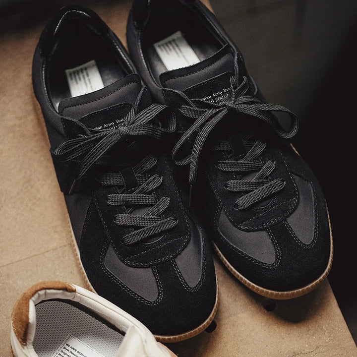 A pair of black lightweight breathable sneakers with premium cow suede and mesh panels, black laces, and gum soles are placed side by side on a brown surface. Part of a white shoe with a beige heel is visible in the lower left corner.
