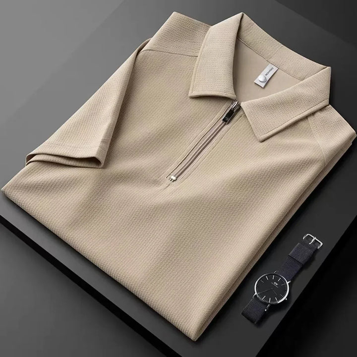 A neatly folded beige men’s silk polo with a front zipper is placed on a dark surface next to a black wristwatch with a minimalist design.