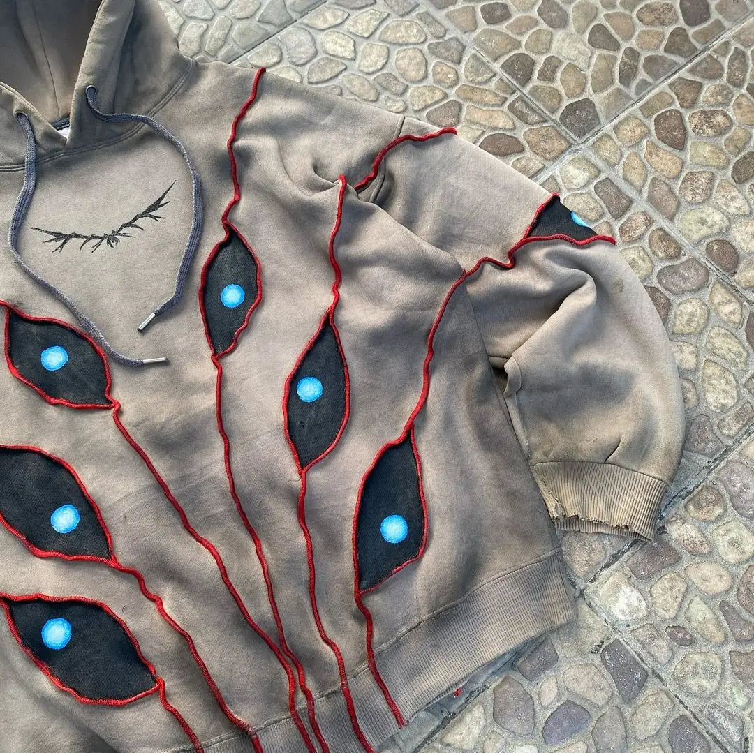 A tan hooded sweatshirt with a comfortable loose fit and red-stitched, abstract eye pattern patchwork featuring blue and black centers, displayed on a stone-tiled surface. The hem and sleeve cuffs are frayed for a distressed look.
