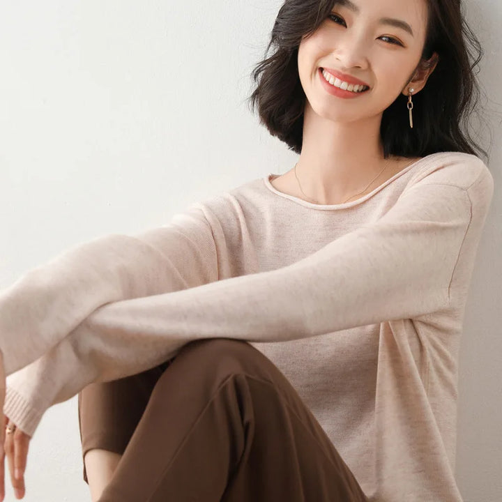 A woman with shoulder-length dark hair, wearing a relaxed fit light beige sweater and brown pants, sits against a white wall, smiling warmly at the camera.