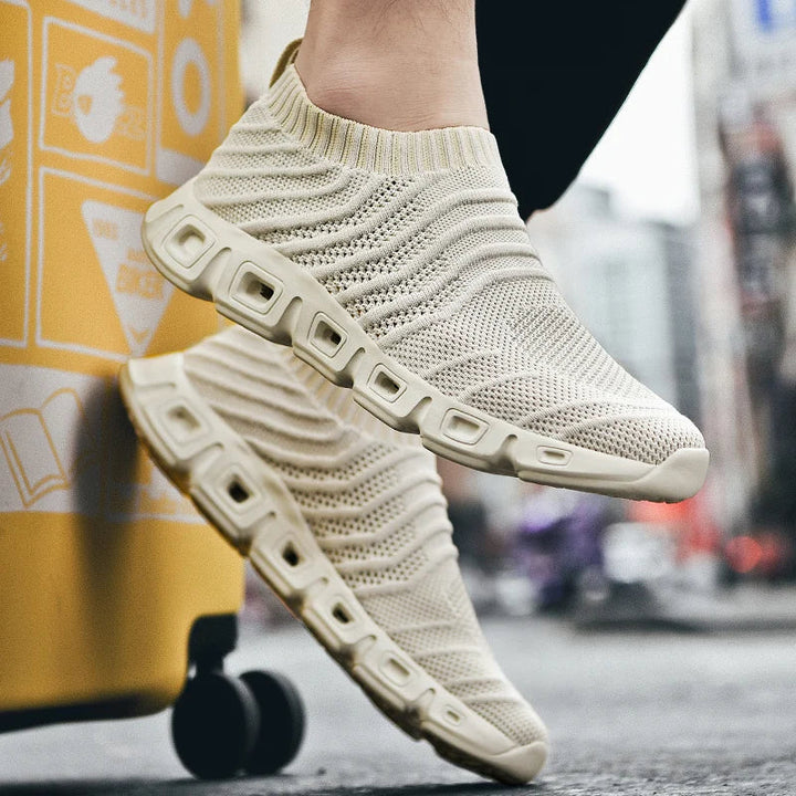 A person wears beige, slip-on knit sneakers with a cushioned sole and rectangular cutouts, offering a sock-like fit while standing outdoors on a city street near a yellow suitcase.