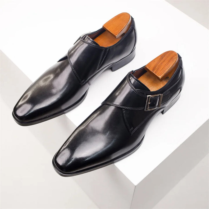 A pair of shiny black Italian leather monk strap shoes with wooden shoe trees, displayed on a white geometric surface.