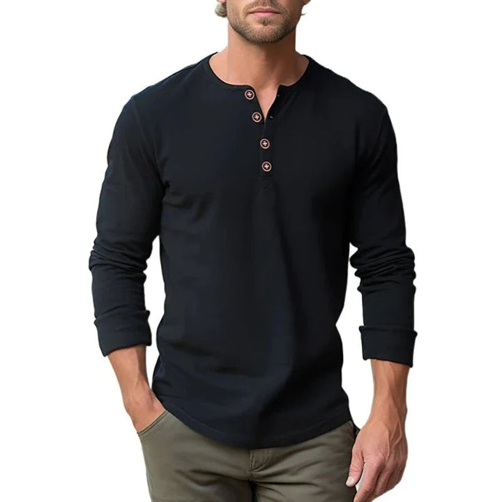 A man wears a classic design, long-sleeve black Henley shirt with four buttons and light brown pants. His hands are in his pockets, and the background is plain white.