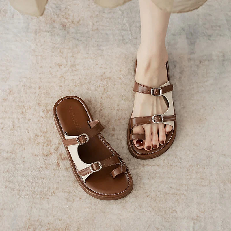 A person wearing brown and white open-toe sandals with a leather upper and dual buckle straps stands on a light textured surface. One sandal is on the foot, while the other with a non-slip rubber sole is placed beside it.
