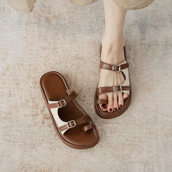 A person wearing brown and white open-toe sandals with a leather upper and dual buckle straps stands on a light textured surface. One sandal is on the foot, while the other with a non-slip rubber sole is placed beside it.