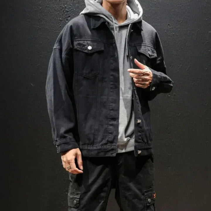 A person wearing a gray hoodie layered under a black premium denim jacket and black pants stands against a dark background. Their face is not visible, and they have tattoos on their fingers.