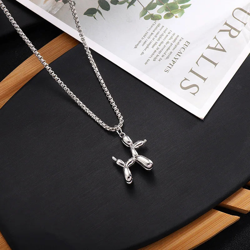 A playful jewelry piece, this silver balloon dog necklace with an adjustable chain rests on a black surface next to a bamboo tray and a magazine.