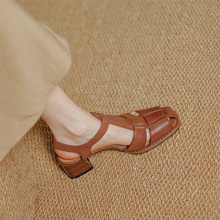 A person wearing tan pants and brown leather, closed-toe, block-heeled sandals handcrafted from Genuine Italian Leather sits on a textured beige carpet.