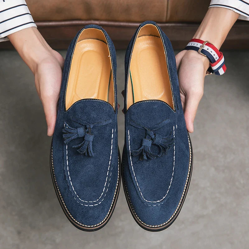 A person holding a pair of blue suede tassel loafers with cushioned footbeds and white stitching, seen from above. The shoes have tan insoles and are held over a gray floor.