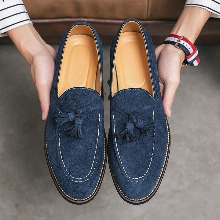A person holding a pair of blue suede tassel loafers with cushioned footbeds and white stitching, seen from above. The shoes have tan insoles and are held over a gray floor.
