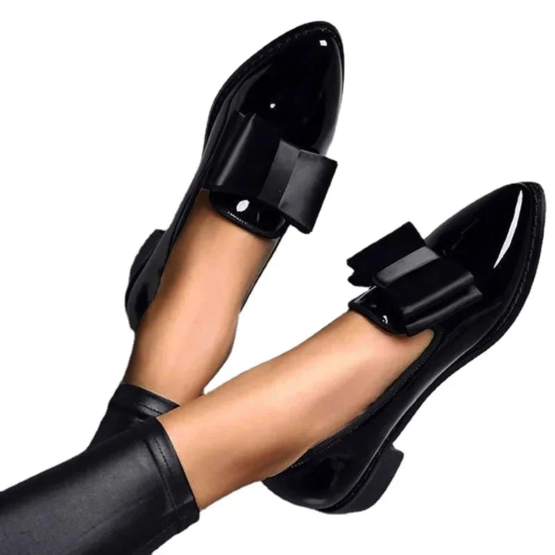 A pair of feet wearing shiny black patent leather loafers with a statement bow on top, shown with black leggings against a plain white background.