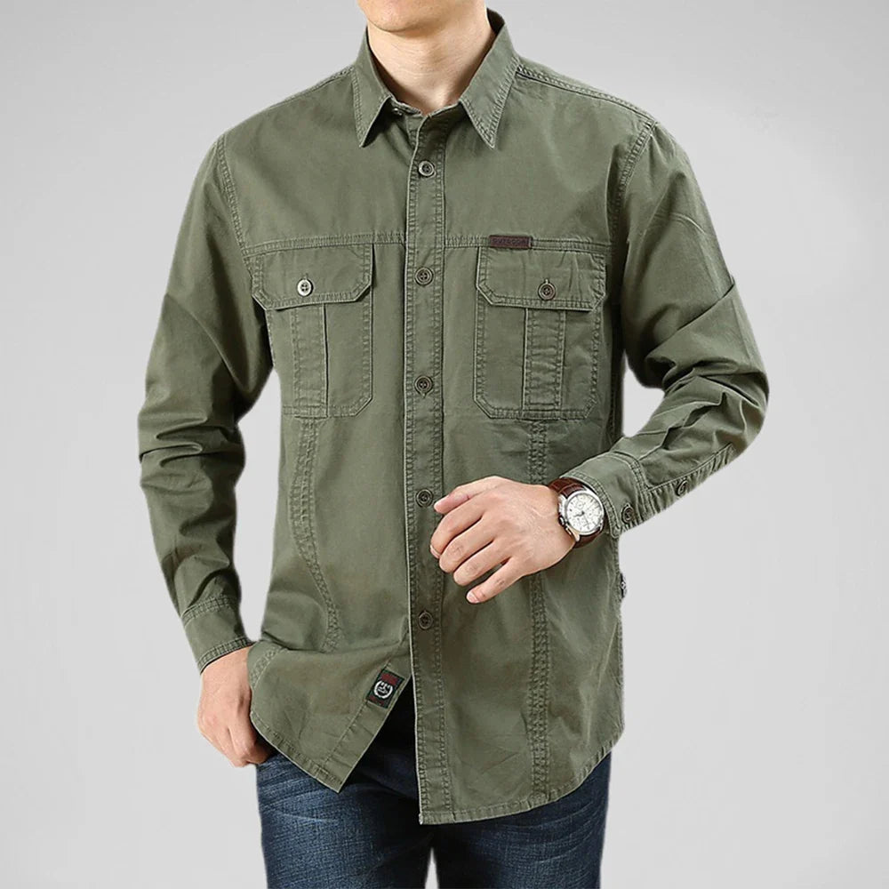 A man wearing a relaxed fit, long-sleeve olive green 100% cotton shirt with two front chest pockets and blue jeans. He poses with one hand in his pocket and the other adjusting his watch against a plain light gray background.