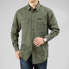 A man wearing a relaxed fit, long-sleeve olive green 100% cotton shirt with two front chest pockets and blue jeans. He poses with one hand in his pocket and the other adjusting his watch against a plain light gray background.