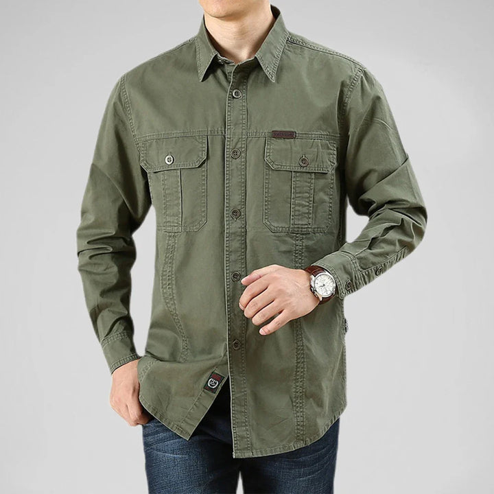 A man wearing a relaxed fit, long-sleeve olive green 100% cotton shirt with two front chest pockets and blue jeans. He poses with one hand in his pocket and the other adjusting his watch against a plain light gray background.