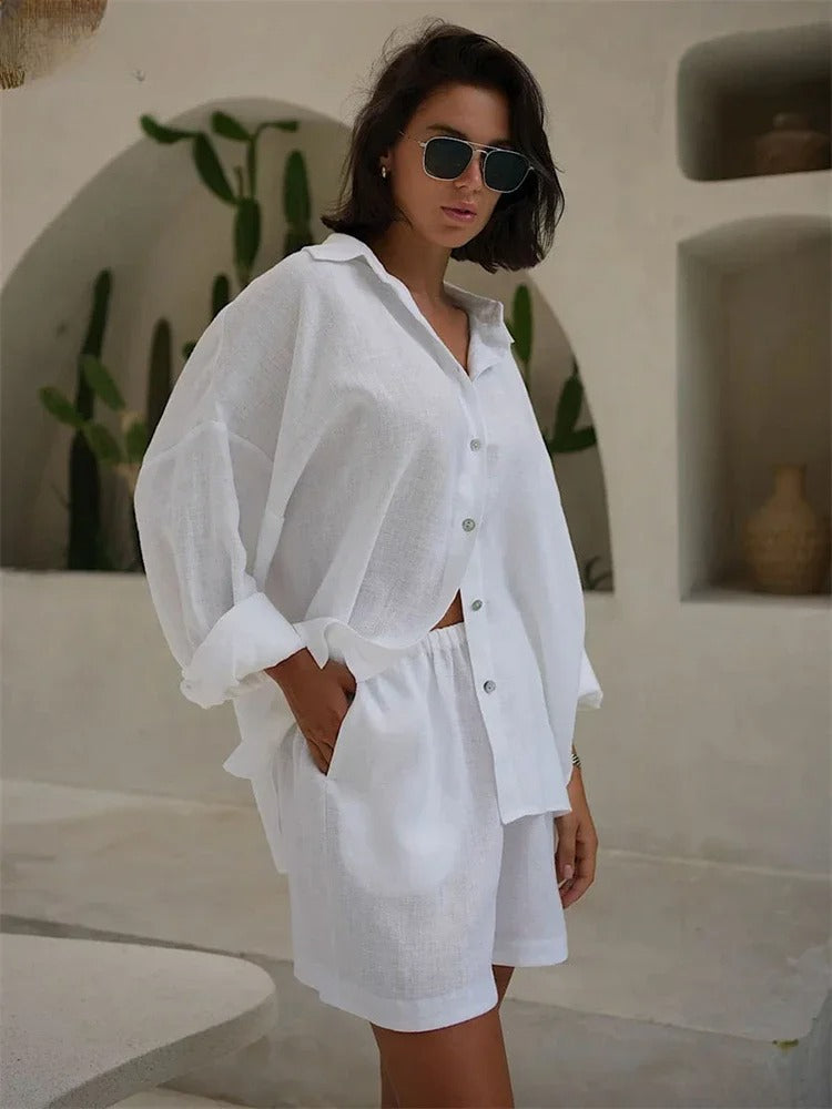 A woman wearing sunglasses and a loose white button-up 2 piece set with matching shorts stands indoors near a wall decorated with green cacti and pottery, embodying effortless summer loungewear style.
