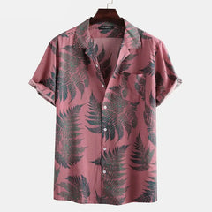 A short-sleeved, button-up cotton shirt with a pink background and a pattern of dark green fern leaves, hanging on a wooden hanger against a plain white backdrop—a classic design perfect for a casual brunch.