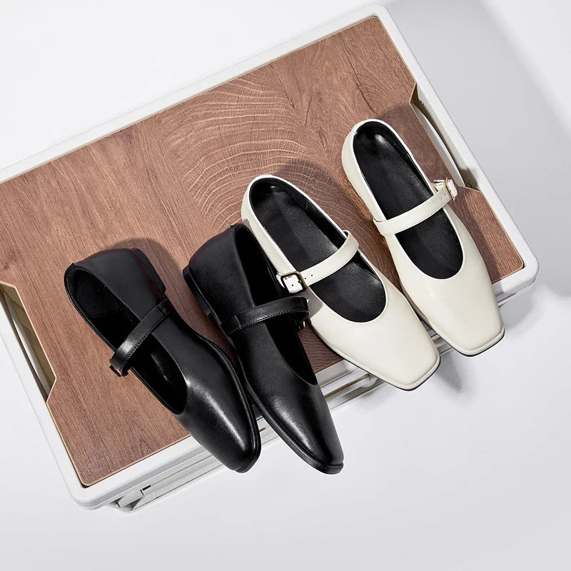 Two pairs of Augusta Leather Ballet Flats, one black and one white, are displayed side by side on a wooden surface with a light background. These handmade leather flats each feature a single strap and a small buckle.