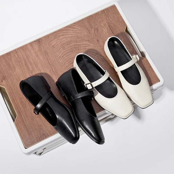 Two pairs of Augusta Leather Ballet Flats, one black and one white, are displayed side by side on a wooden surface with a light background. These handmade leather flats each feature a single strap and a small buckle.
