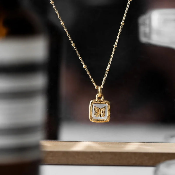 A gold necklace with a rectangular pendant featuring a butterfly design showcases elegant craftsmanship, displayed against a blurred background with black, white, and brown tones.